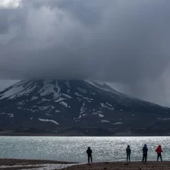 Visitantes em pé na Laguna del Diamante sob nuvens tempestuosas que cobrem o vulcão Maipo.
